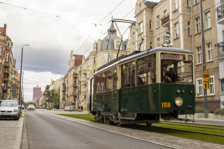 Na zdjęciu widnieje ciemnozielony tramwaj na torowisku, w tle kamienice