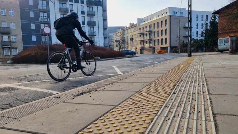 Nowa platforma przystankowa z matami naprowadzającymi i ostrzegawczymi, w tle skwer, na jezdni rowerzysta