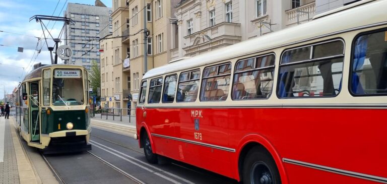 Dwa mijające się pojazdy - na wprost stary, ciemnozielony tramwaj a obok niego czerwony autobus