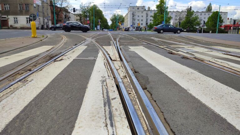 Przejście dla pieszych przez torowisko. Widoczne szyby i spękany, pofałdowany asfalt