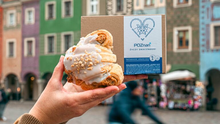 Trzymany w ręku rogal na tle kartonu z napisem Poznań życzy smacznego i kolorowych kamienic