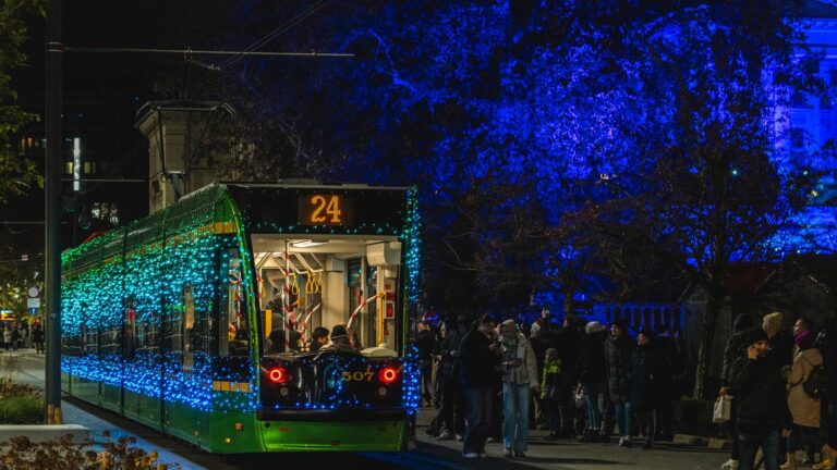 Oświetlony lampkami tramwaj na przystanku. Obok niego tłum osób